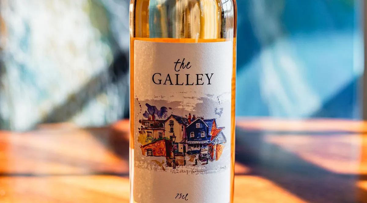 A bottle of The Galley rosé wine with its watercolour Topsham label, lit by the sun on a wooden table