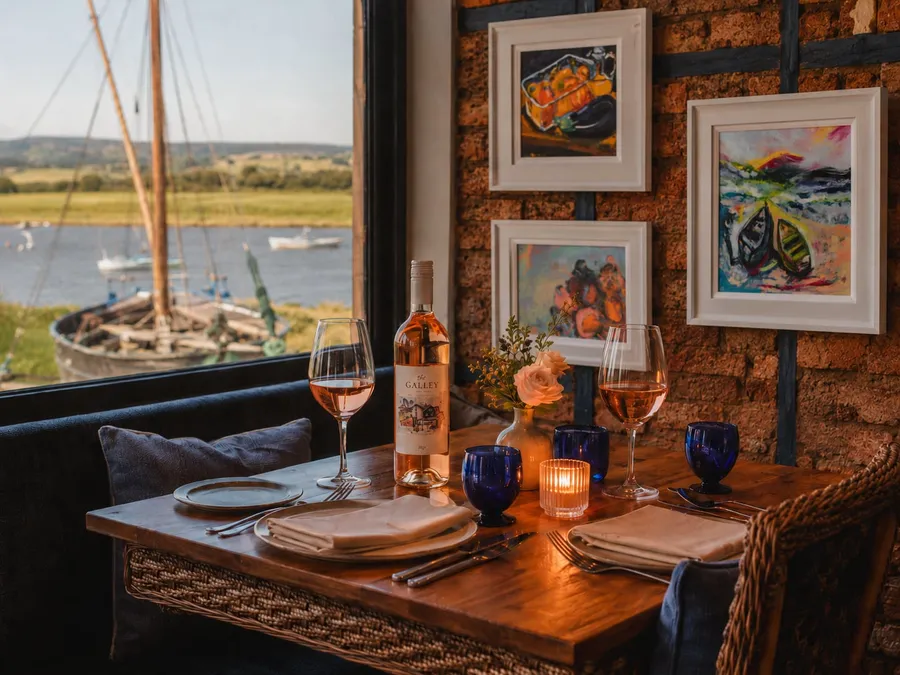 A candlelit table for two by the window at The Galley, Topsham — rosé wine and cobalt glasses, framed coastal art on the brick wall, and a sailing boat on the Exe Estuary beyond at golden hour
